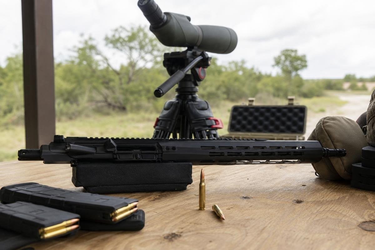 A rifle upper receiver, magazines, and ammunition on a wooden shooting bench with a spotting scope on a tripod behind them.