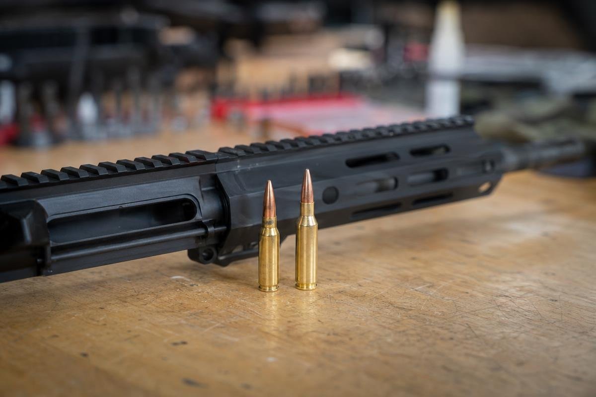 An AR upper receiver with M-LOK handguard on a wooden workbench with a 6mm ARC and a larger cartridge standing upright in front of it.