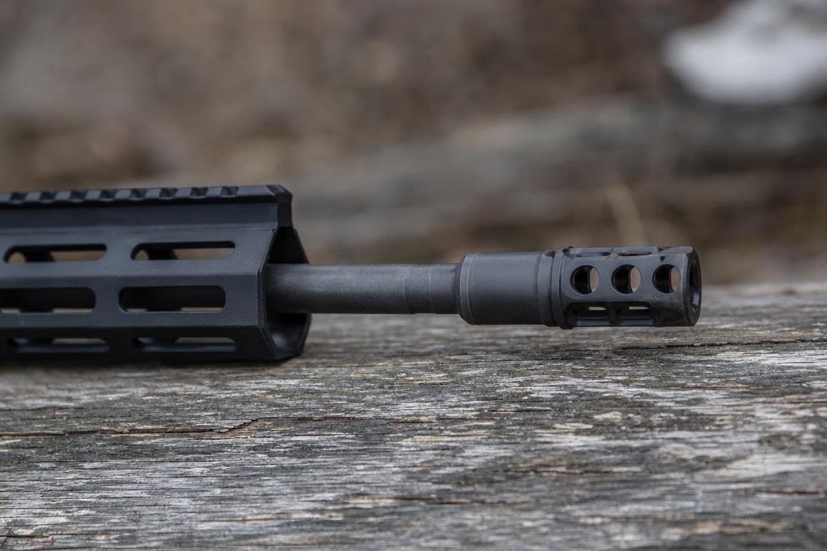 Close-up of a rifle barrel with a muzzle brake installed, resting on a wooden surface.