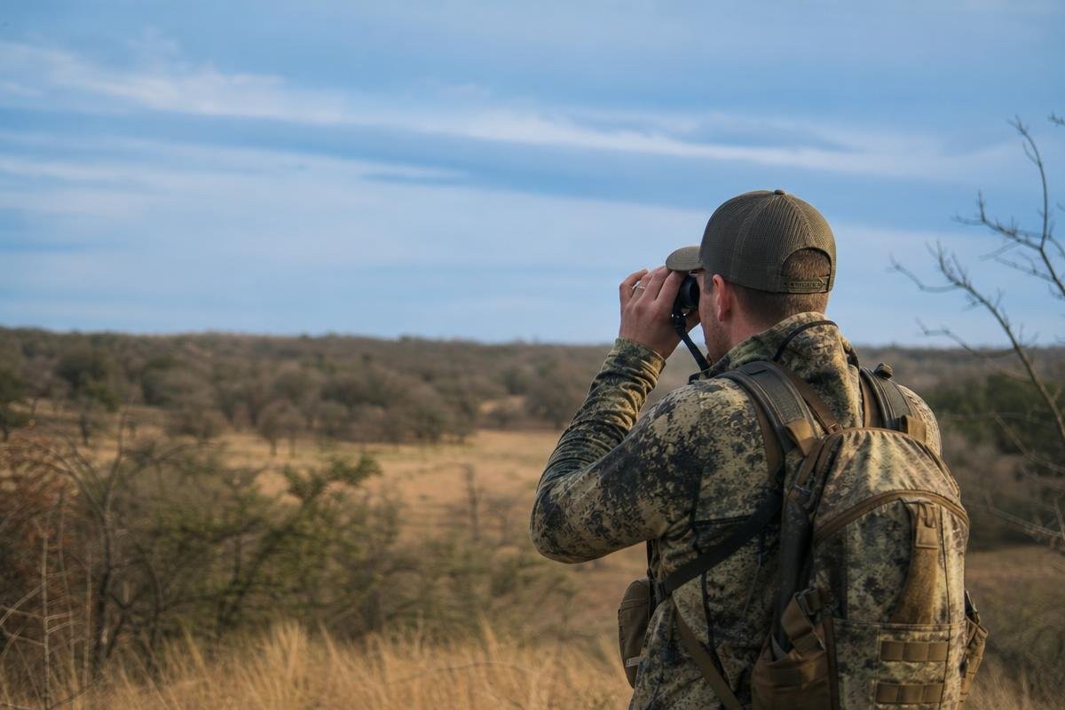 A hunter in camouflage looking through binoculars across a field of dry grass and trees.