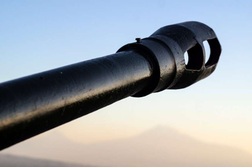 A close-up view of a rifle barrel with a muzzle device against a blurred background.