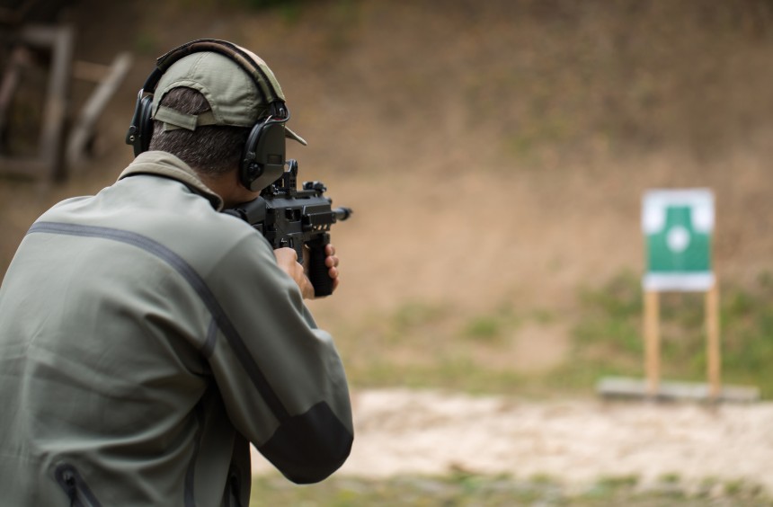 A shooter aiming a scoped rifle at a distant target on an outdoor range.