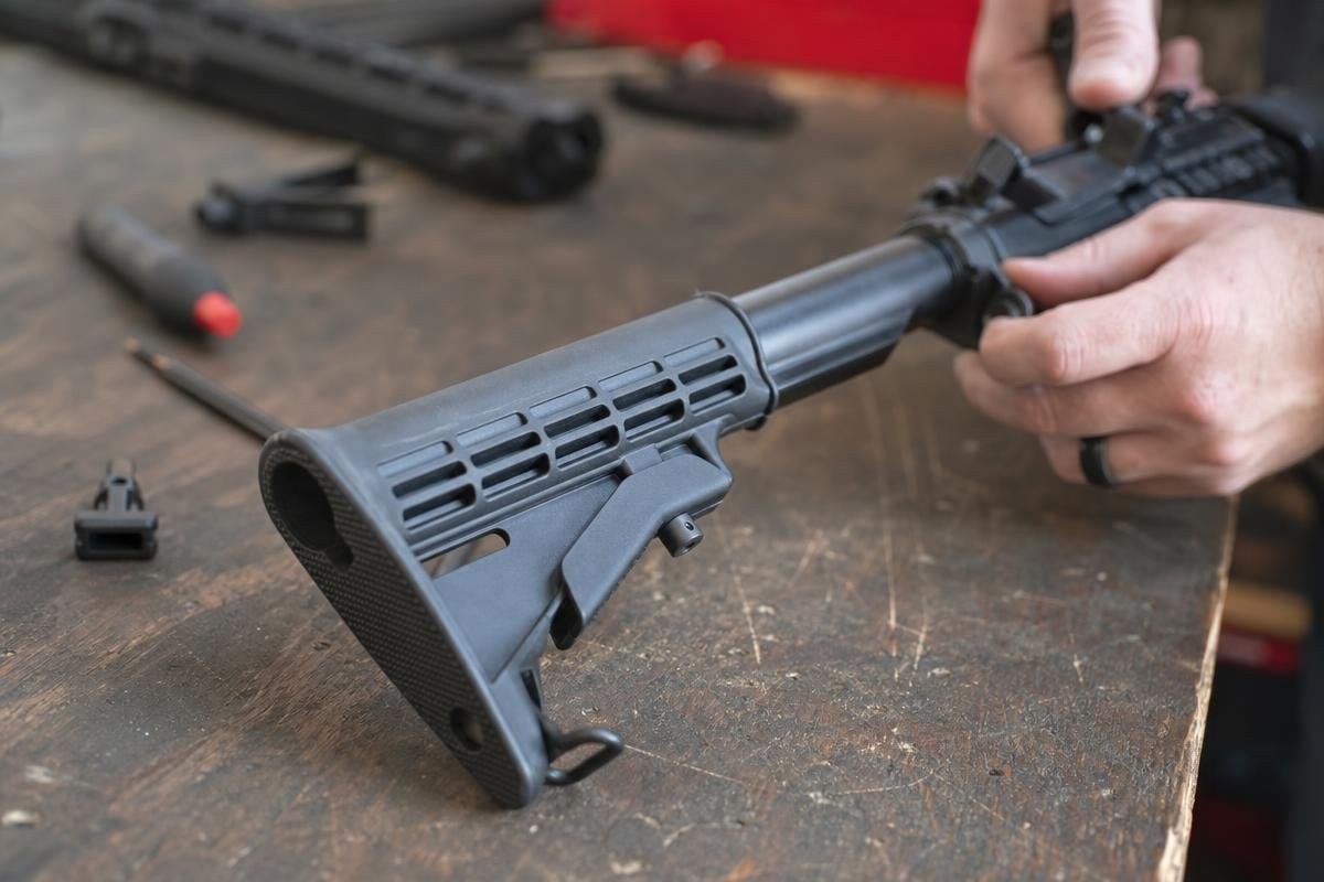 A pair of hands installing a collapsible stock onto an AR-15 buffer tube on a workbench.
