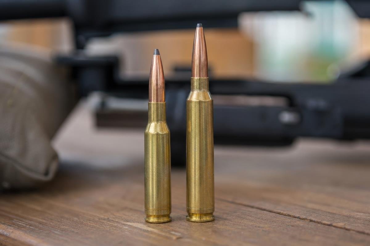 Two large rifle cartridges standing upright on a wooden bench with rifle components blurred in the background.
