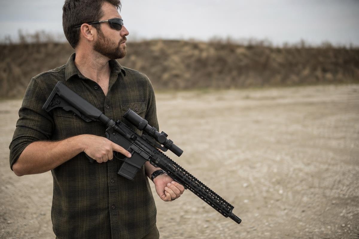 A shooter in a green flannel shirt holding an AR-15 rifle fitted with a low power variable optic.