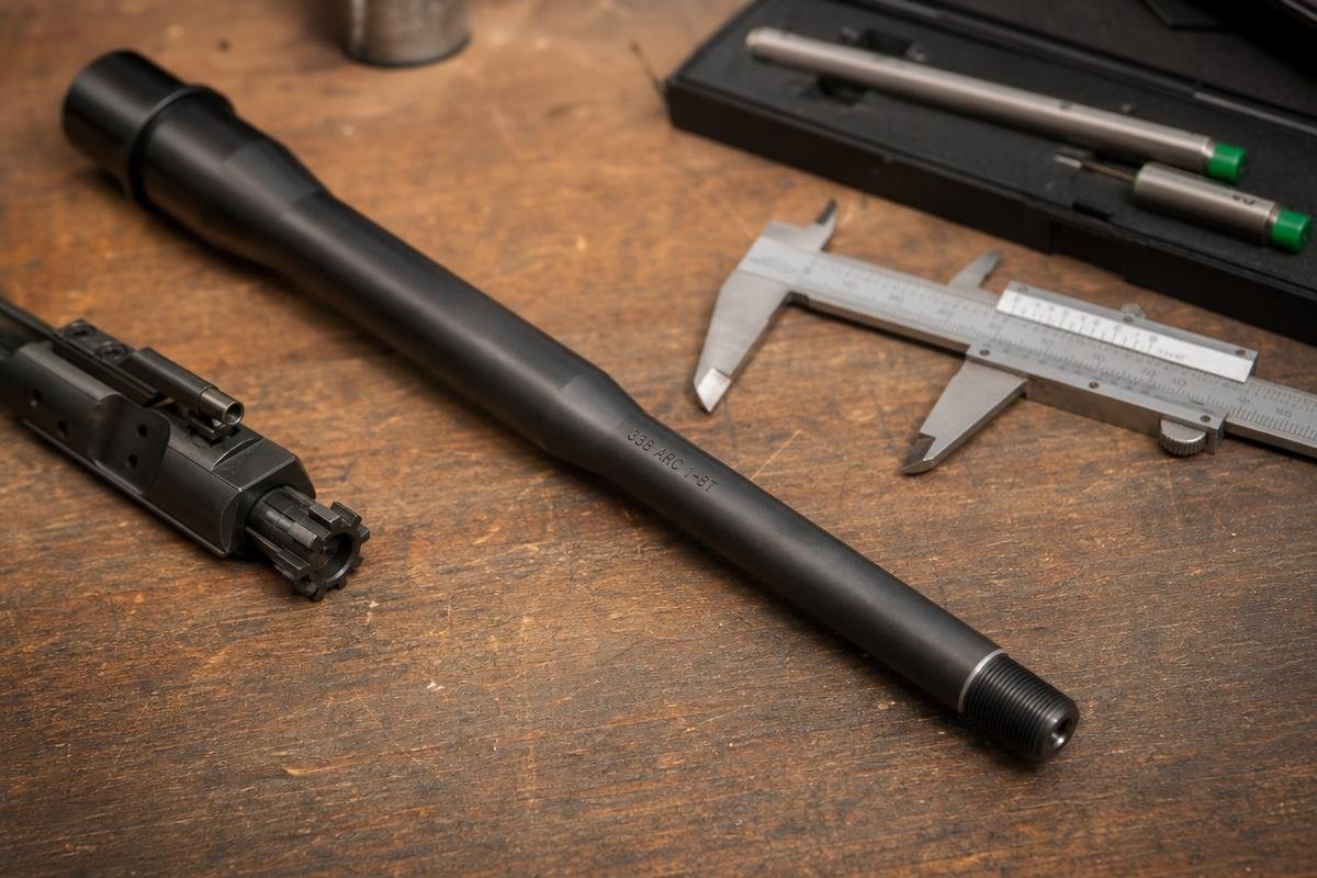 A 338 ARC barrel resting on a workbench next to a bolt carrier group and a pair of calipers.