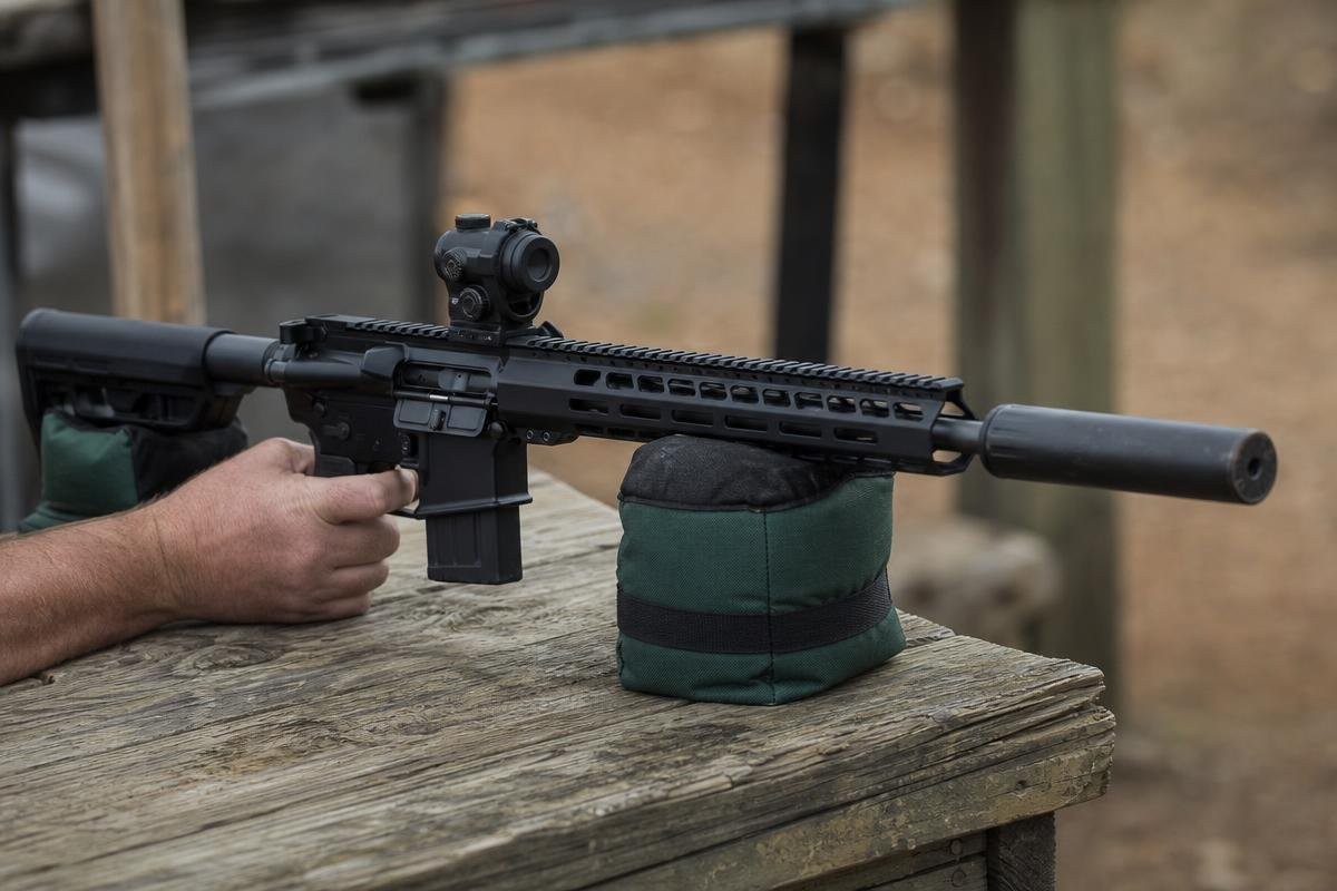 A person holding a suppressed rifle on a shooting rest.