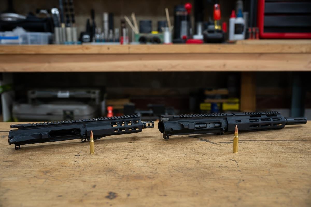 Two black AR upper receivers with M-LOK handguards on a wooden workbench, each with a rifle cartridge standing upright in front of it.