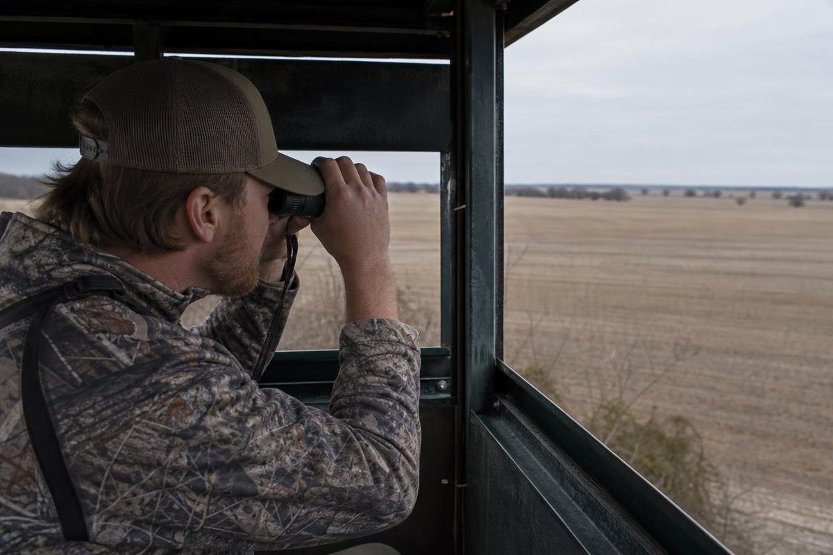 A hunter looking through binoculars from inside a hunting blind overlooking a field.