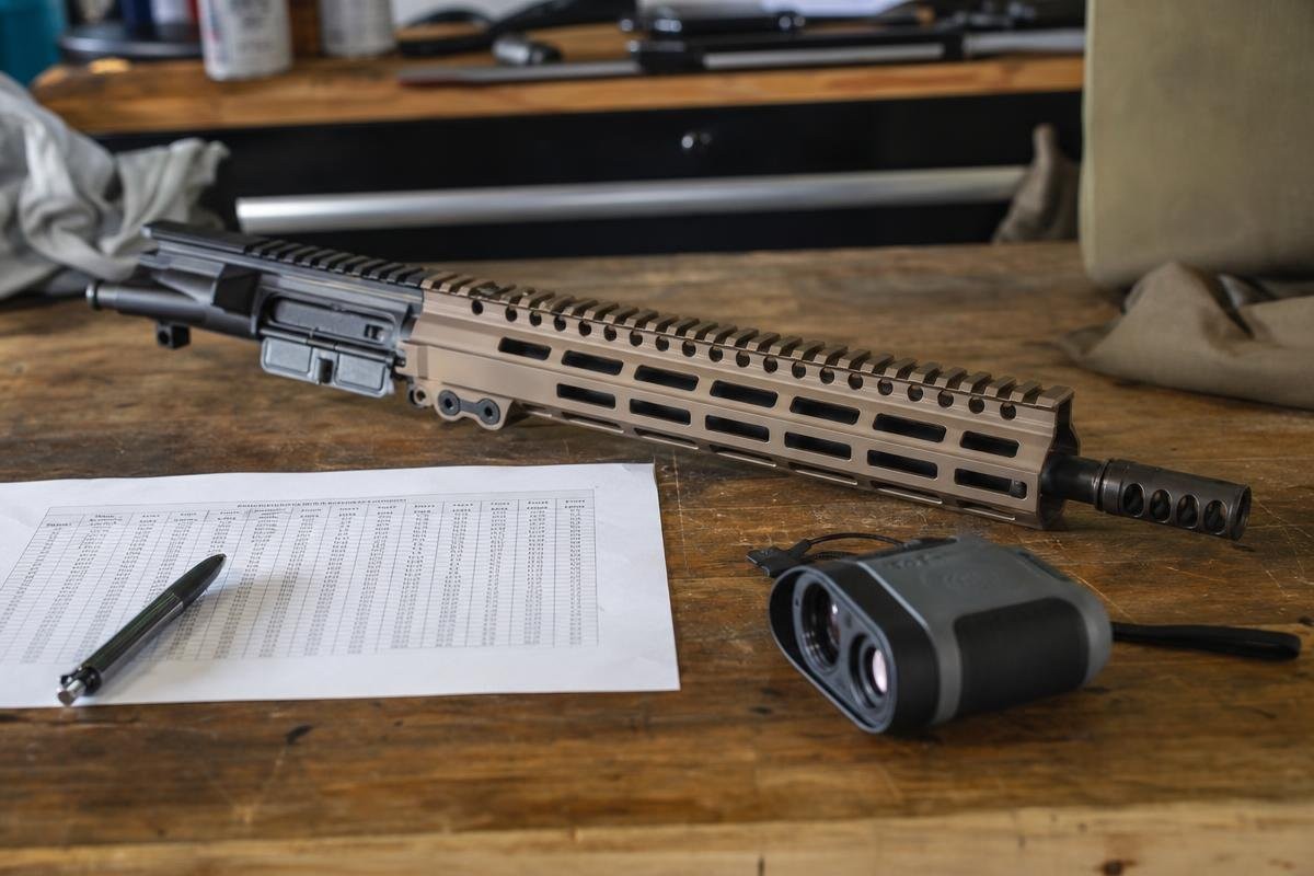 A 338 ARC upper receiver assembly on a wooden workbench next to a printed ballistics chart, a pen, and a rangefinder.