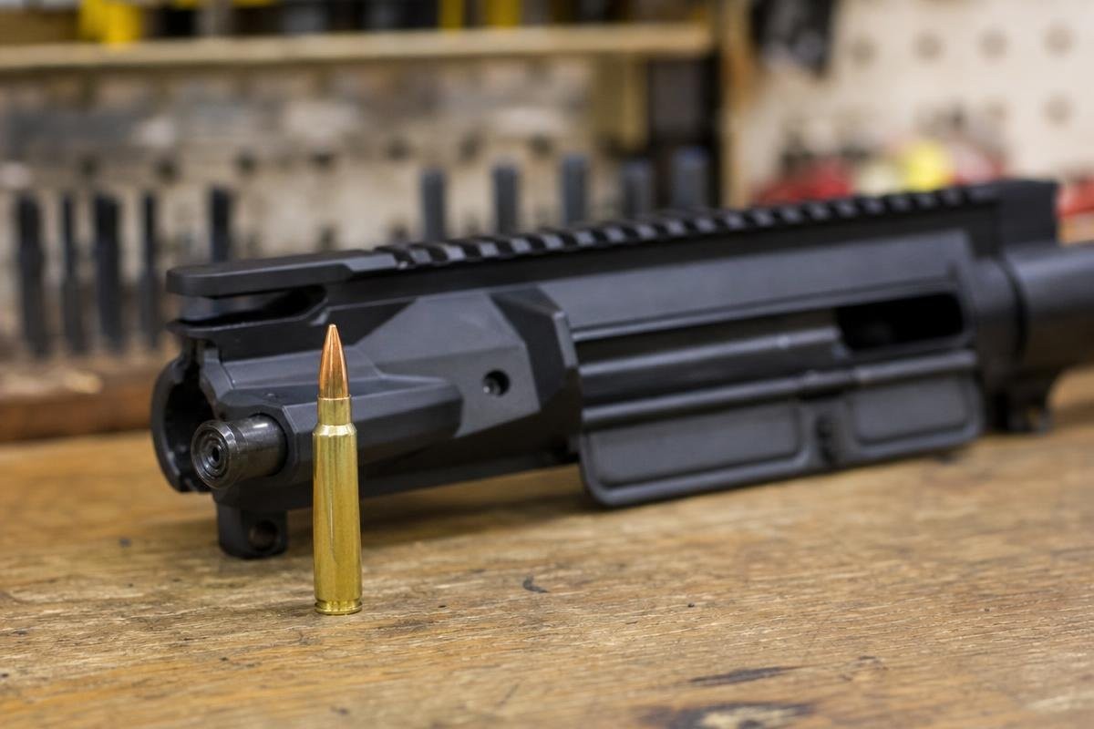 A 338 ARC cartridge standing upright in front of an AR-15 upper receiver.