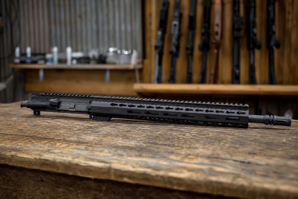 A black AR-15 upper receiver assembly resting on a wooden workbench in front of a wall rack of rifles.