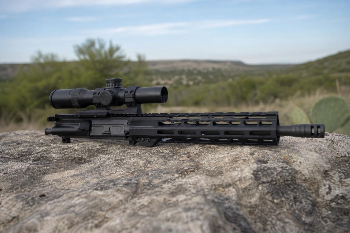 A 6.5 Creedmoor AR-platform upper receiver with a mounted scope resting on a rock in an open field.