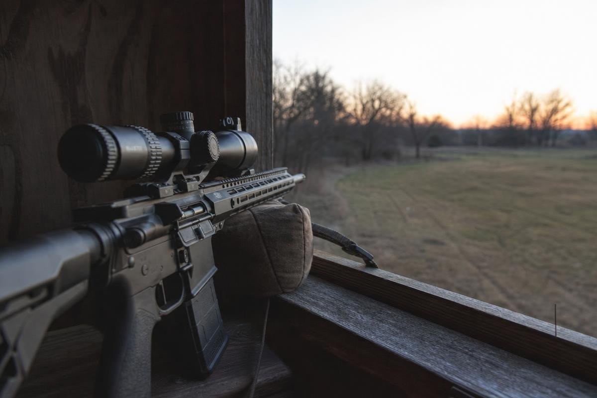 A rifle with a scope resting on a shooting rest in a deer blind overlooking a field at sunset.
