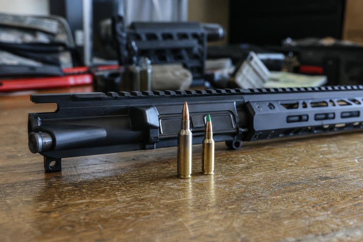 An AR-15 upper receiver on a wooden table with a 338 ARC cartridge and a 5.56 cartridge standing in front of it.