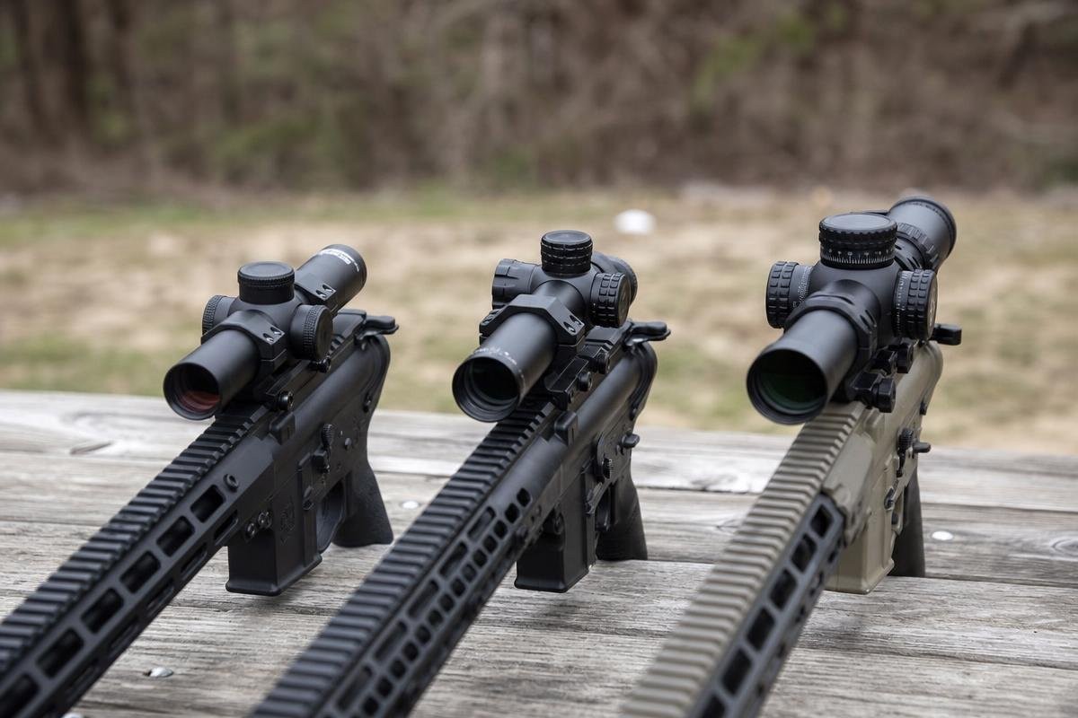 Three AR rifles with low power variable optics mounted, resting on a wooden surface.