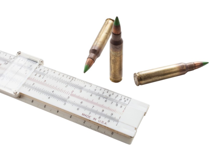 Two 338 ARC cartridges standing upright behind a slide rule on a workbench.
