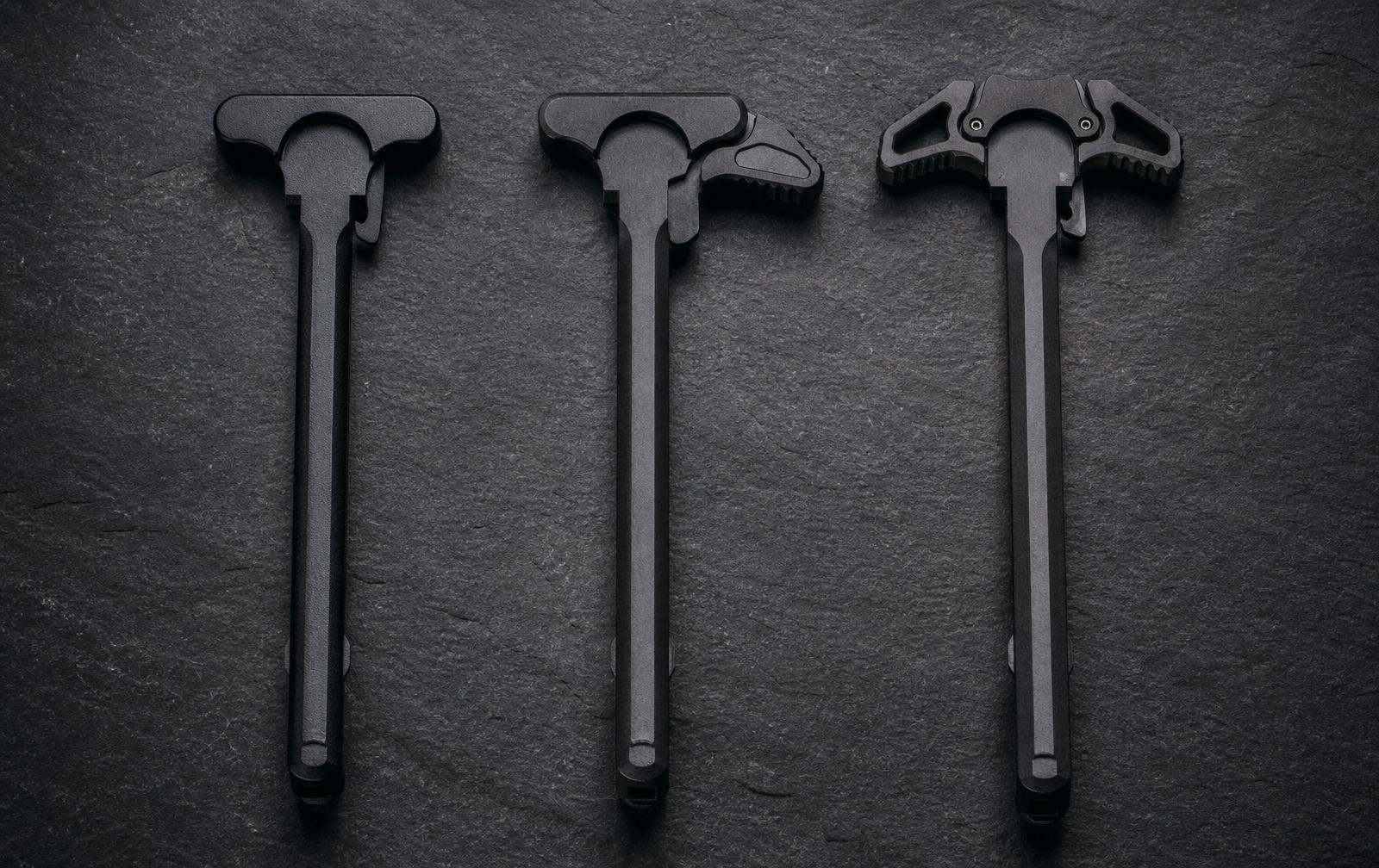 Three AR-15 charging handles side by side: standard mil-spec, extended tactical, and ambidextrous, on dark slate.