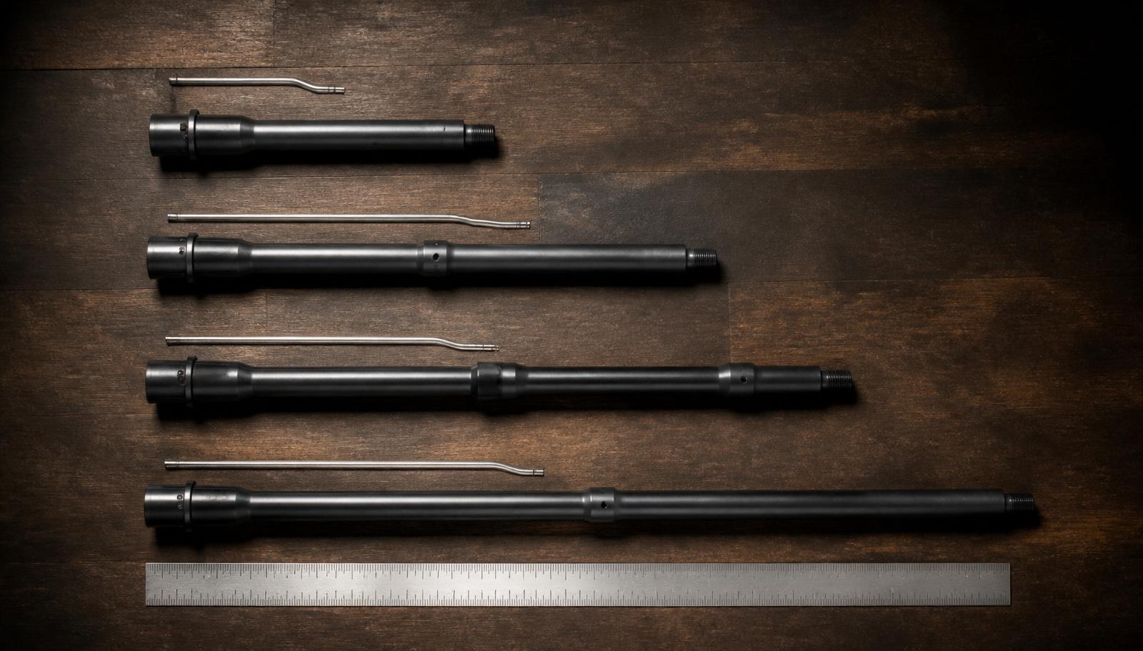 Overhead flat lay of four AR-15 barrels and their gas tubes on dark walnut, showing pistol, carbine, mid-length, and rifle gas system lengths.