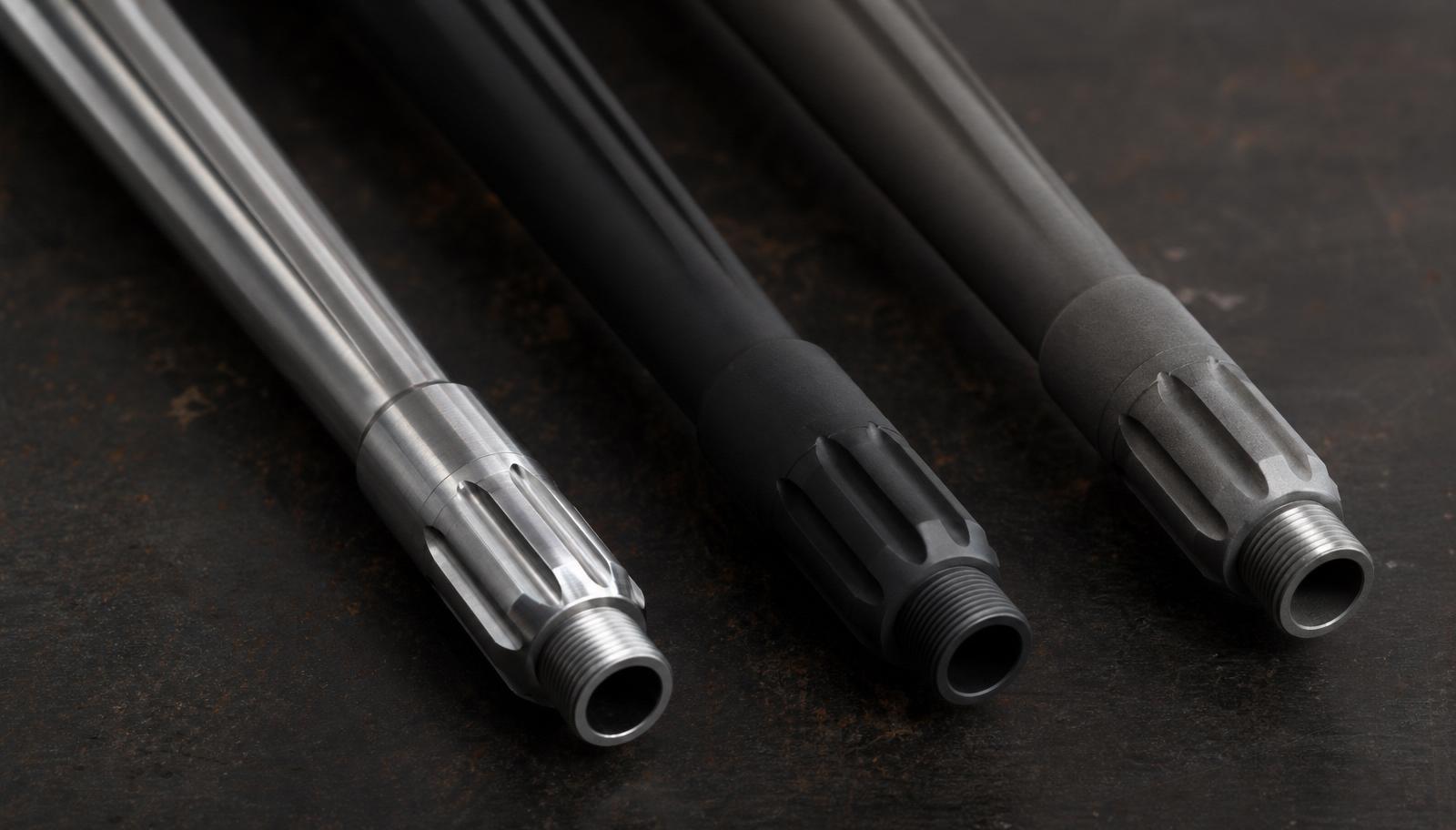 Three AR-15 barrels laid side by side on a dark surface: polished stainless, matte black nitride, and grey chrome-lined.
