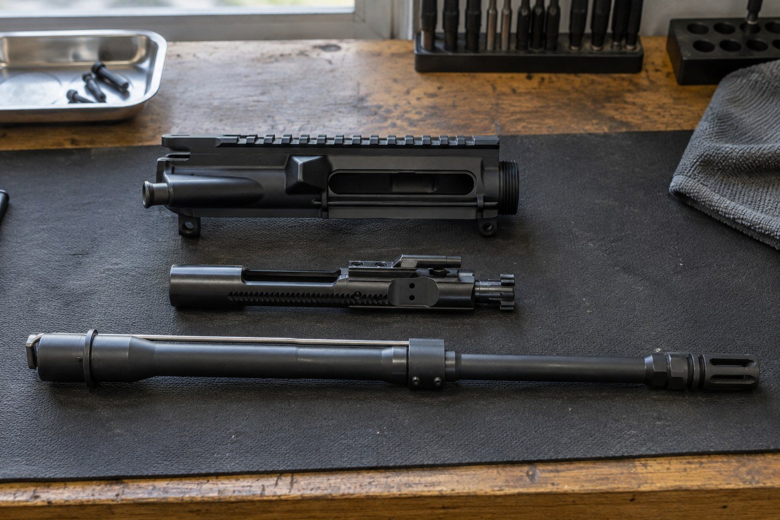 Disassembled black rifle components arranged on a dark work mat beside tools and a parts tray